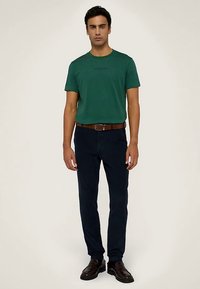 Zelené bavlněné tričko s kulatým výstřihem a logem, v kombinaci s tmavě modrými kalhotami a hnědým pásem, stojící na neutrálním pozadí.