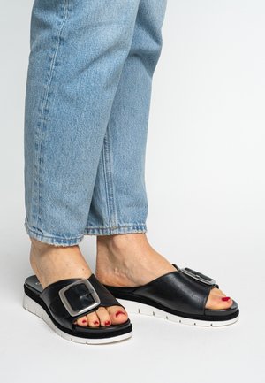 Sandalias tipo slide de cuero negro con una gran hebilla plateada, plantilla contorneada, suela blanca y diseño de punta abierta, combinadas con jeans de mezclilla deshilachados.