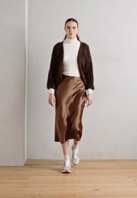 Brauner, texturierter Cardigan über einem weißen Rollkragenpullover, dazu ein fließender brauner Satinrock. Kombiniert mit weißen Sneakers und sichtbaren weißen Socken.