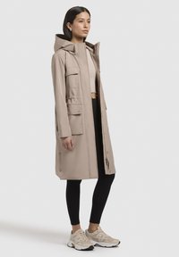 Beige vízálló parka kapucnival, elülső zsebekkel, cipzáros záródással és egyenes szabással. Fekete leggings fölött és bézs sportcipővel viselve.