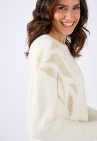 Suéter color crema con una textura suave y esponjosa que presenta un patrón geométrico dorado. Cuello redondo, mangas largas, corte relajado.