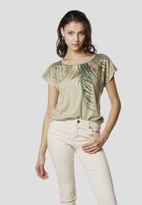 Gaiši zaļa īsroku t-krekls ar palmās lapas grafiku tumšākos zaļumos un metāliskām akcentēm, kombinēts ar krēmkrāsas pieguļošiem džinsiem.
