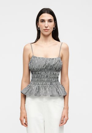 Kvinde med langt mørkt hår iført sort og hvid ternet smocket ærmeløs top med flæsekant og hvide bukser mod en ensfarvet baggrund.