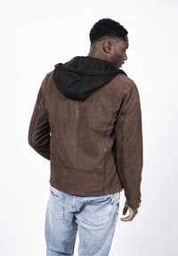 Brun mocka jacka med en svart huva, som visar upp en avslappnad design och synliga sömnadsdetaljer. Bärs med ljusblå denimjeans.