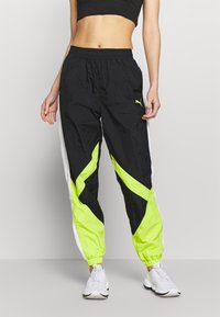 Slim-fit black track pants with neon yellow and white color blocks, elastic ankles, a small Puma logo, worn with white sneakers.