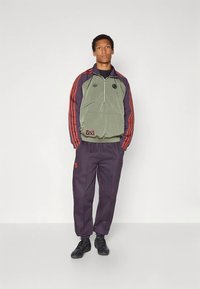 Πράσινο και μοβ μπουφάν με φερμουάρ και κόκκινες ρίγες, σε συνδυασμό με μοβ παντελόνι με κόκκινες λεπτομέρειες και μαύρα παπούτσια. Ομαλή υφή σε όλο το ένδυμα.