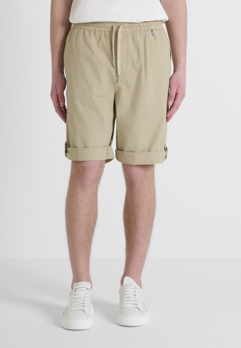 Shorts in cotone beige con vita elastica e cordoncino, caratterizzati da orli arrotolati e tasche laterali. Abbinati a sneakers bianche.