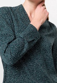 Blouse à manches longues en tissu vert foncé avec un motif marbré de pois blancs et bleus clairs. Elle présente un décolleté en V et des manches à boutons.