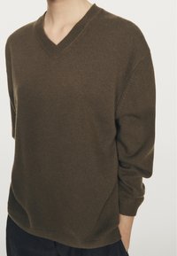 Persona che indossa un maglione a V marrone a maglia con maniche lunghe e pantaloni scuri, in piedi contro uno sfondo chiaro e uniforme.