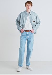 Cămașă cu dungi albastre și albe, tricou alb dedesubt, blugi albaștri deschisi, centură maro și pantofi albi. Poziție în picioare pe un fundal simplu.