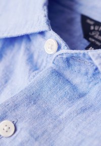 Camisa de tejido azul claro con textura y botones blancos, primer plano de un botón con el texto "Superdry".