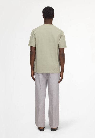 Homme vu de dos portant un t-shirt beige clair à manches courtes et un pantalon ample gris à fines rayures avec des chaussures noires sur fond blanc.