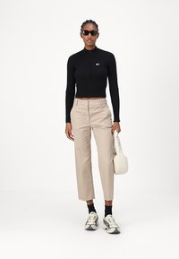 Melna ribota cropped top ar rāvējslēdzēju, béža piegrieztas bikses, melnas zeķes, baltas masīvas kedas un maza balta soma ar logo detaļu.