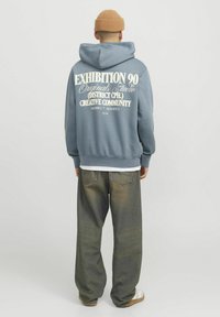 Sudadera azul con un gran texto gráfico blanco en la parte de atrás, combinada con unos vaqueros verdes anchos y zapatillas crema. Gorro de punto en la cabeza.