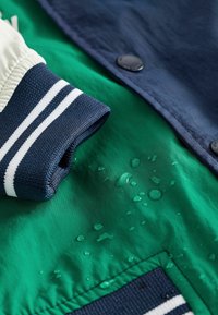 Veste imperméable verte et marine avec poignets élastiques, rayures blanches, boutons-pression, et gouttes d'eau sur la surface du tissu.