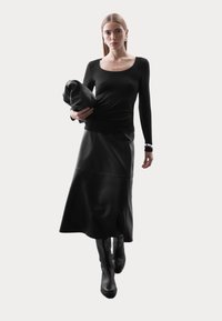 Zwarte langemouwen top, getailleerd in de taille, gecombineerd met een zwarte leren rok. Het model houdt een zwart kledingstuk vast, draagt kniehoog laarzen en zilveren oorbellen.