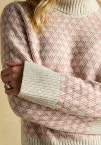 Nahaufnahme von gekreuzten Armen, die einen cremefarbenen und rosa gemusterten Strickpullover mit gerippten Bündchen tragen, und einem silbernen Ring an der linken Hand.