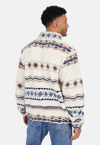 Uomo che indossa una giacca in pile bianca con motivi geometrici blu, marroni e beige, abbinata a jeans blu, girato di spalle su uno sfondo semplice.