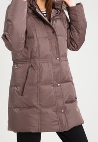 Brun puffjacka med huva som har en fuskpälsdetalj, dragkedjefickor på sidorna, tryckknappsstängning och quiltat mönster.