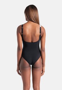 Μαύρο ολόσωμο μαγιό με ρυθμιζόμενους ιμάντες, σχεδιασμό με χαμηλή πλάτη και διαφανείς λεπτομέρειες από δίχτυ κατά μήκος των πλευρών. Ομαλή υφή υφάσματος.