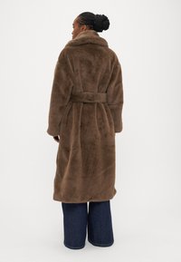 Hnedý kabát z umelého kožušiny s vysokým golierom, opasok v páse a dlhými rukávmi, siahajúci do polovice lýtok, nosený cez široké rifle s rovnými nohavicami.