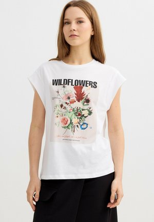 Mujer con camiseta blanca sin mangas con texto "WILDFLOWERS" y diseño floral colorido, combinada con pantalones negros sobre fondo blanco.