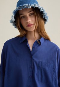 Camisa azul de botones con un bolsillo, hecha de una tela suave; combinada con un sombrero de cubo de denim deshilachado en un tono de azul más claro.