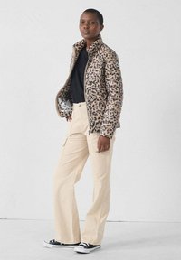 Chaqueta acolchada con estampado de leopardo en tonos beige y marrón, cierre frontal con cremallera, combinada con pantalones cargo beige y zapatillas negras.