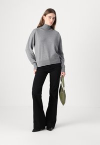 Sweater à col roulé gris avec une coupe décontractée, ourlet côtelé, associé à un pantalon noir évasé. Modèle tenant un sac à main vert. Fond simple.