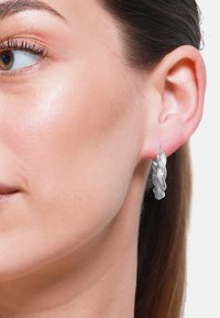 Strieborný skrútený kruhový náušník vystavený na uchu modelu, predvádzajúci textúrovaný, pletený dizajn. Náušník má lesklý kovový povrch.