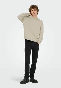 Lichtbeige trui met een ronde halslijn, gecombineerd met zwarte jeans. Het model staat met één hand op zijn nek en draagt zwarte schoenen.