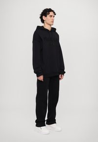Felpa nera con tasca frontale e design grafico testurizzato, abbinata a pantaloni neri e sneakers bianche. Silhouette moderna e semplice.