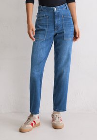 Personne portant un jean en denim bleu avec de grandes poches avant et des baskets beige avec des rayures rouges, se tenant contre un mur uni.