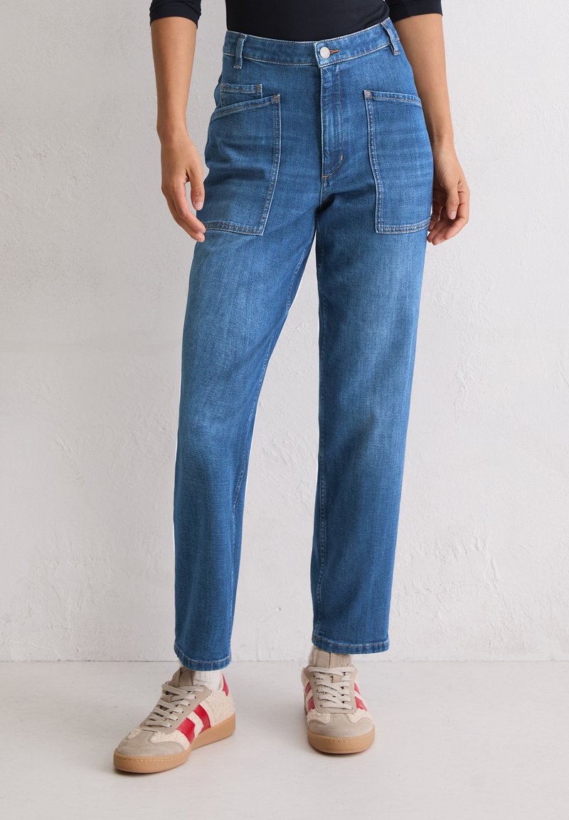 Personne portant un jean en denim bleu avec de grandes poches avant et des baskets beige avec des rayures rouges, se tenant contre un mur uni.