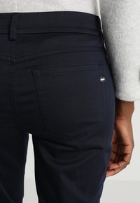 Navy modré džíny s přiléhavým střihem, které mají zadní kapsu s dekorativním stehem a jemný vícebarevný pruh na okraji kapsy.