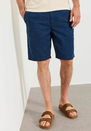 Mann trägt marineblaue gemusterte Shorts und braune Sandalen mit Schnallen und steht auf grauem Boden vor einer weißen Wand.