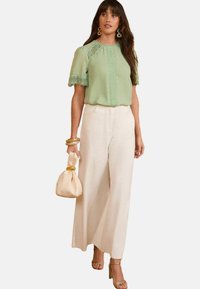 Top verde chiaro con dettagli in pizzo abbinato a pantaloni beige a vita alta e a gamba larga. Gli accessori includono una borsa beige e bracciali dorati.