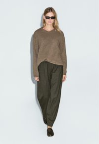 Maglione di lana marrone con scollatura a V, abbinato a pantaloni larghi verde oliva e mocassini neri. Il modello indossa occhiali da sole neri. Sfondo bianco.