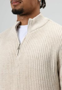 Man draagt een roomkleurige, gebreide trui met halve rits en een wit T-shirt eronder, gecropt net onder de neus en boven de borst.