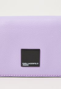 Sviestinės tekstūruotos odos piniginė, turinti juodą kvadratinį logotipo pleistrą su užrašu "KARL LAGERFELD JEANS" baltu tekstu, papildyta rožinėmis siūlėmis.
