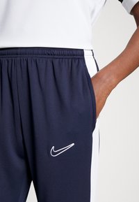 Pantaloni jogger blu navy con pannelli laterali bianchi, vita elastica e logo Nike ricamato in bianco. Il materiale è liscio, leggero e sportivo.