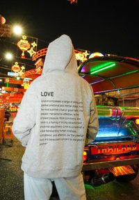Un sweat à capuche gris avec un grand texte imprimé au dos détaillant le concept de l'amour. Porté avec un pantalon blanc ample ; lumières néon colorées en arrière-plan.