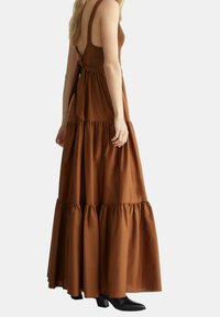 Robe maxi marron avec un corsage en crochet ajusté, un dos ouvert et une jupe à volants. Le tissu semble lisse avec une silhouette fluide. Bottines noires jusqu'à la cheville.