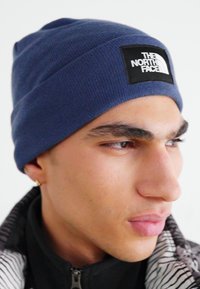 Blå stickad mössa med en åtsittande design, prydd med en svart fyrkantig etikett med varumärket "The North Face" i vit text.