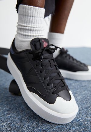 Osoba nosząca białe skarpetki i czarne sneakersy z grubymi białymi podeszwami stoi na niebieskim dywanie.