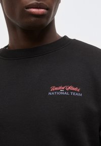 Κοντινό πλάνο ενός ατόμου που φοράει μαύρο φούτερ με το κείμενο «United States Men's National Team» σε κόκκινο και μπλε στο στήθος.