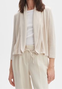 Beżowy, lekki kardigan z otwartym przodem, szerokimi rękawami i teksturowanym splotem. Zestawiony z białą bluzką i jasnymi spodniami.