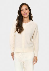 Femme avec de longs cheveux bruns portant un cardigan crème boutonné et un pantalon de couleur claire, posant devant un fond blanc uni.