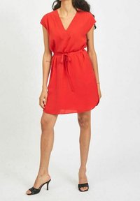 Robe rouge courte avec un décolleté en V, des manches courtes, une taille cintrée avec un lien et un tissu fluide. Portée avec des talons ouverts noirs.