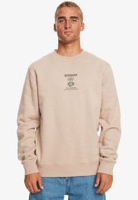 Sweatshirt beige clair avec un col rond, arborant un imprimé graphique gris foncé à l'avant et des poignets côtelés aux manches et à l'ourlet.